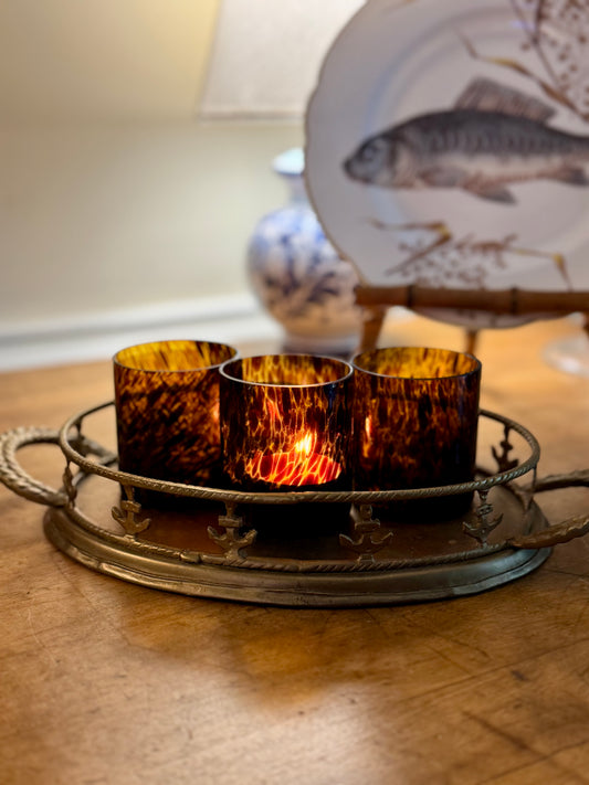 Three amber-colored candles in a decorative metal holder on a wooden surface with a fish-themed plate in the background.