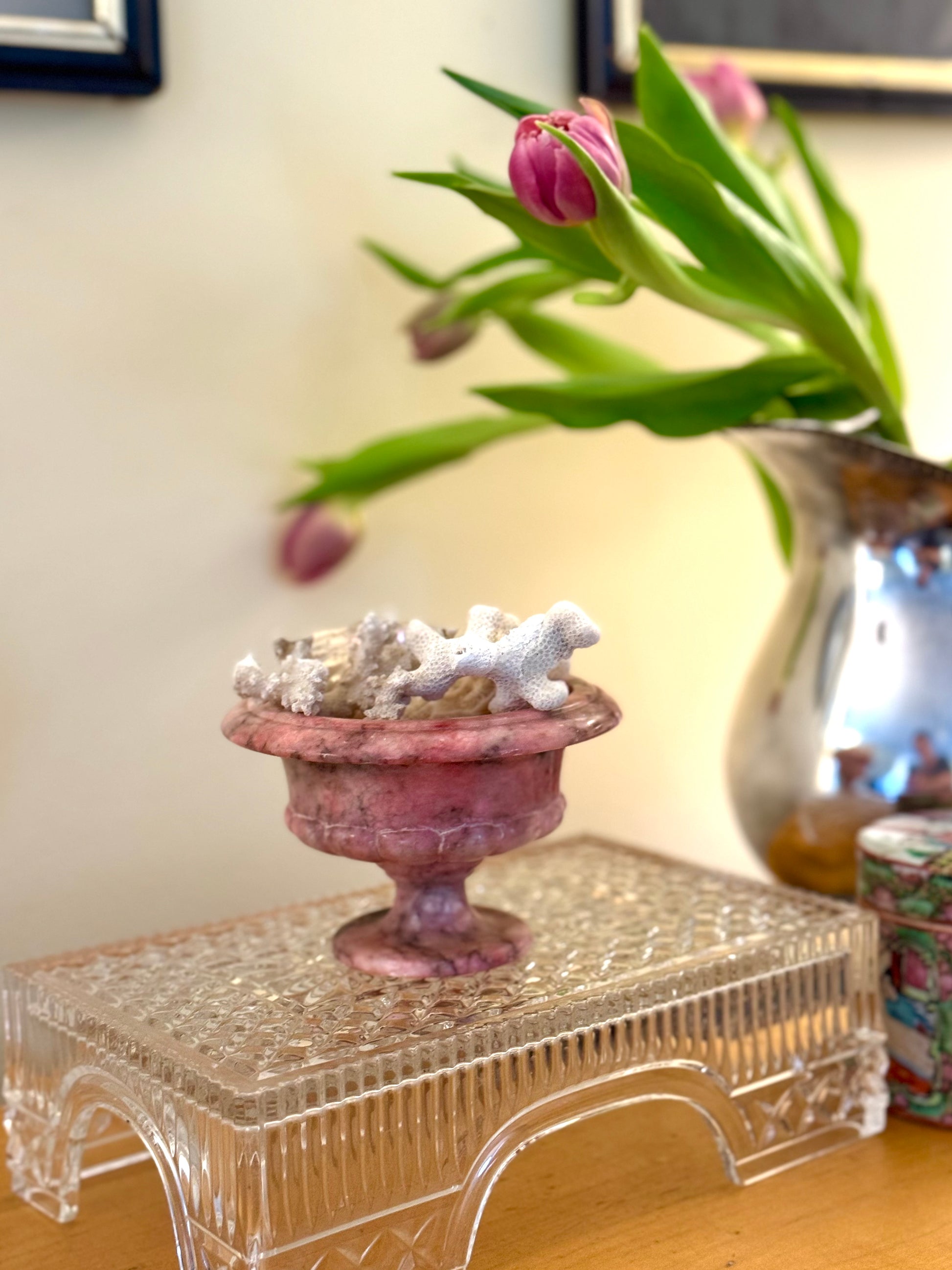 Decorative pink urn with seashells on a crystal stand, next to a vase with pink tulips.